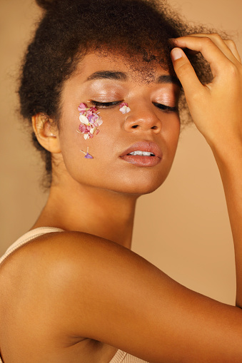 Portrait of beautiful African girl with curly hair shows creative make-up with flower petals while hand gently touches her face isolated over beige background. Natural eco cosmetics and beauty concept