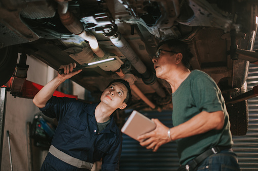 Asian Chinese senior mechanic and trainee checking car undercarriage in auto repair shop