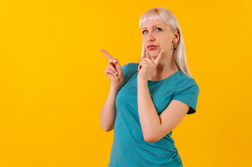 Portrait with pensive pointing posture, blonde caucasian girl in studio on yellow background