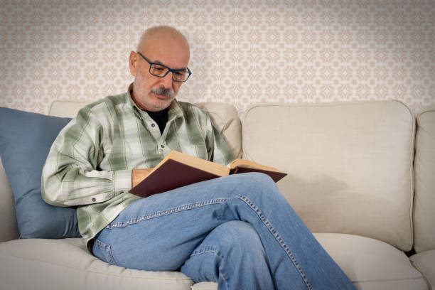 istock Happy Senior Man Sitting on Sofa and Reading Book at Home 1449619040