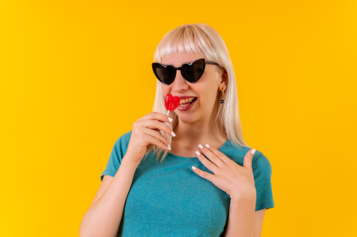 Having fun eating a heart lollipop, blonde caucasian girl on yellow background studio