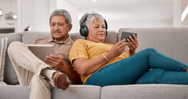 couple senior, détendez-vous avec la technologie et la tablette, le smartphone et les écouteurs, le streaming et le visionnage en ligne avec le réseau 5g. personnes âgées, homme et femme avec appareil, canapé et ensemble à la retraite. - senior couple audio photos et images de collection