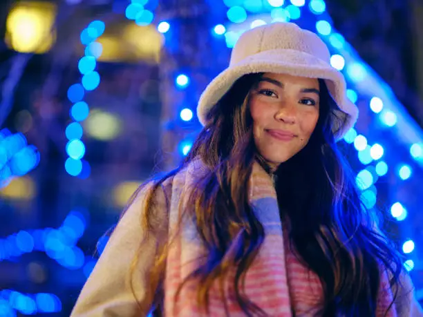 A young woman in a downtown area of a city in the evening, with holiday lights in the background.