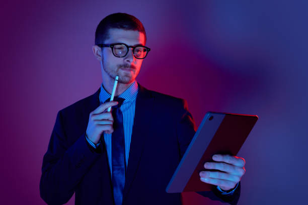 istock Young man, manager wearing business suit and eyeglasses using digital tablet on gradient blue purple background in neon light. Studying, work, job, finance 1449628740
