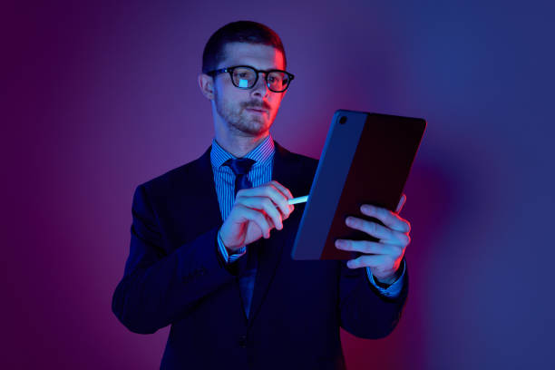 istock Young man, manager wearing business suit and eyeglasses using digital tablet on gradient blue purple background in neon light. Studying, work, job, finance 1449628746