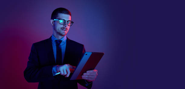 istock Young man, manager wearing business suit and eyeglasses using digital tablet on gradient blue purple background in neon light. Studying, work, job, finance 1449628749