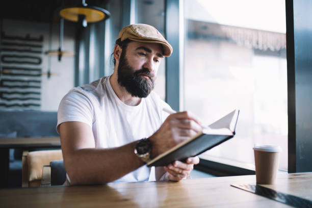 istock Serious bearded mature man in hat sitting in cafe interior writing in notepad during remote job, concentrated 40s male author making notes and planning ideas spending time on research for project 1449630755