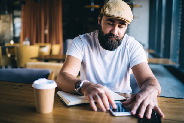 istock Serious bearded male working in cafe interior writing in notebook and making research on smartphone, pensive mature man in trendy hat making booking via application on smartphone connected to 4G 1449630778