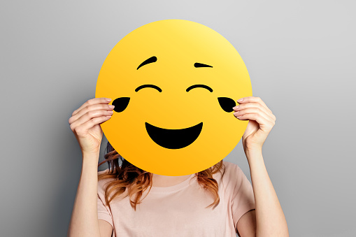 Laughing emoticon with tears of joy. Girl holds a yellow smiley with romantic love face emoticon isolated on a grey studio background