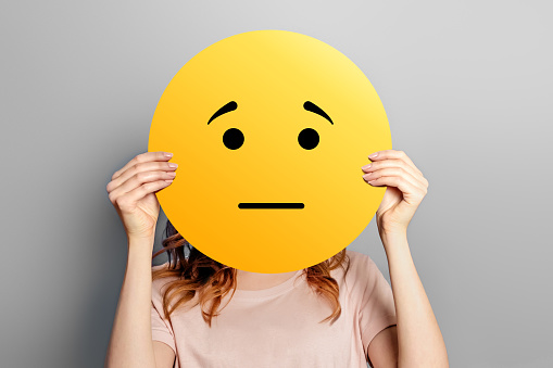 offended emoticon. Girl holds a yellow smiley with sad face isolated on a gray studio background