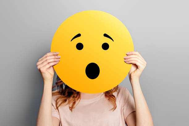 wow schockierte emoji. mädchen hält einen gelben smiley mit überraschtem gesichts-emoticon isoliert auf einem grauen hintergrund. cartoon-gesicht. - smiley face smiling sign people stock-fotos und bilder