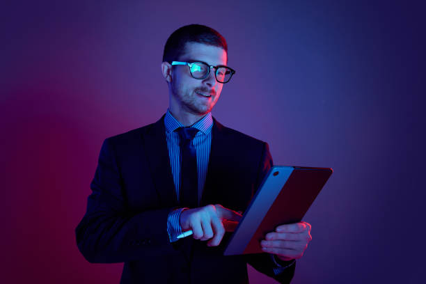 istock Young man, manager wearing business suit and eyeglasses using digital tablet on gradient blue purple background in neon light. Studying, work, job, finance 1449638599