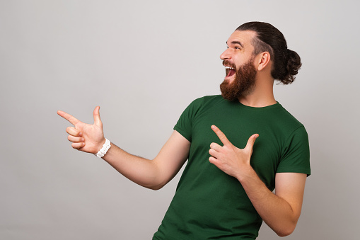 Handsome excited bearded man wearing green is pointing up with two fingers over light grey background.