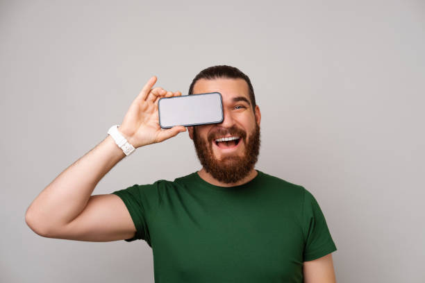 istock Smiling bearded man covers an eye with his phone showing screen for copy space. 1449641948
