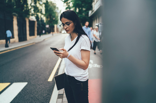 Millennial woman in optical eyewear using internet connection for browsing blog website, Spanish generation Z standing at roadside and checking email message during smartphone online chatting