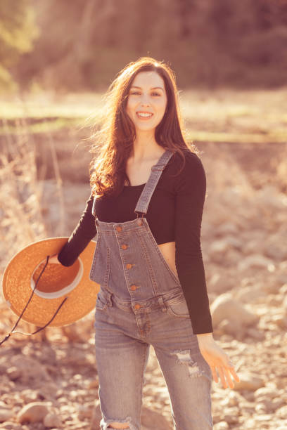Country girl wearing bib overalls smiling and holding straw hat to one side. Photo is heavily backlit