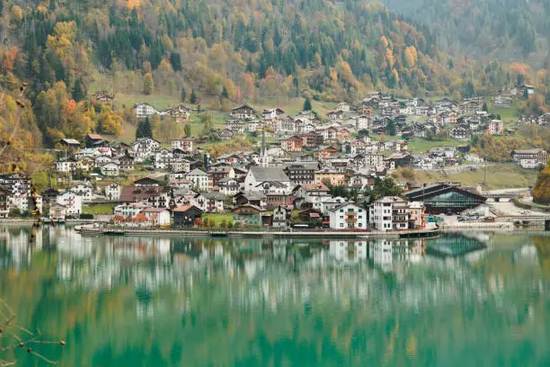 close up view of the town of Alleghe in autumn, extending along the slopes of a mountain, reflecting on the emerald green water surface of its own lake.  Belluno, Veneto region, Italy