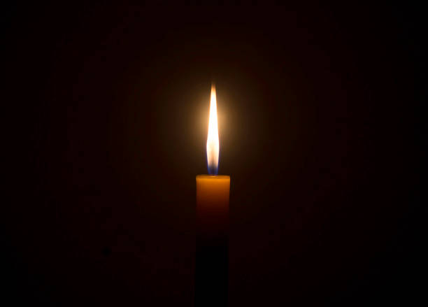 A single burning candle flame or light glowing on an orange candle on black or dark background on table in church for Christmas, funeral or memorial service with copy space