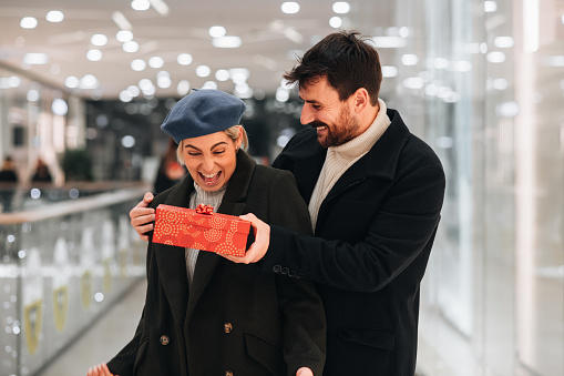 Fashionable couple celebrating Christmas. Couple buying gifts at the mall. Man holding hand over girlfriend’s eyes, surprising her with a gift. Christmas is the season of giving.