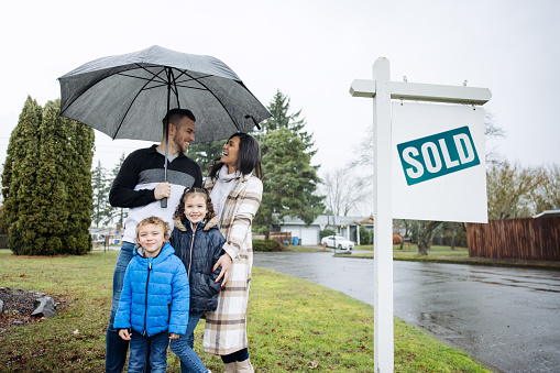 A young Caucasian family smiles, celebrating the purchase of a new home in the Pacific Northwest.  Typical Washington rain falls on the scene, the father holding an umbrella.