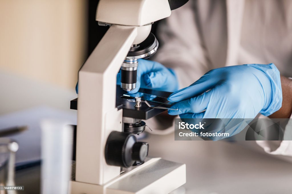 Lab tech in protective gloves putting a slide with a scientific sample under a microscope Close up shot of unrecognizable laboratory technician, in blue disposable protective gloves, putting a slide with a scientific sample under a microscope to analyze. DNA Stock Photo