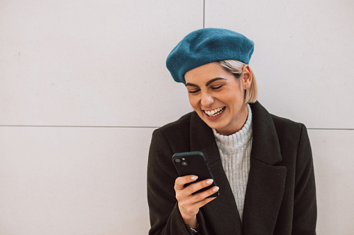 Beautiful young woman with blonde hair using her smart phone, laughing, having a good time while texting with her friends. Cute girl with a beret and Winter clothes standing in a shopping mall alone.