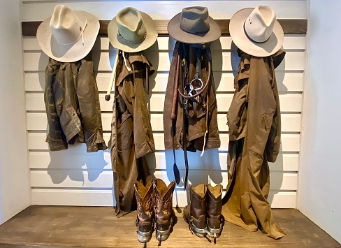 Horizontal still life of country style cowboy boots with horse riding spurs, riding jackets and hats with bridle on wood wall hook and bench  on rural farm horse property Bangalow NSW Australia