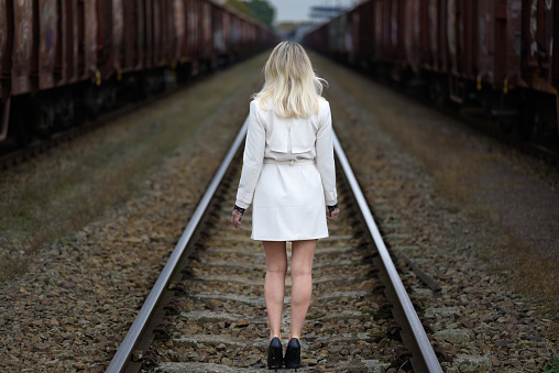 Rear view of a middle-aged attractive blond woman in a white trench coat standing on train tracks in front of train wagons