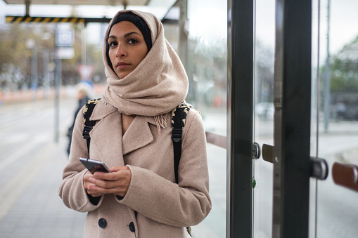 Young muslim woman with a smartphone waiting for bus at city bus stop.