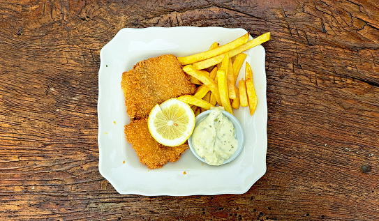 Deep fried fish and chips with lemon, french fries, and tartar dipping sauce, served on a white plate, on a wooden rustic background.