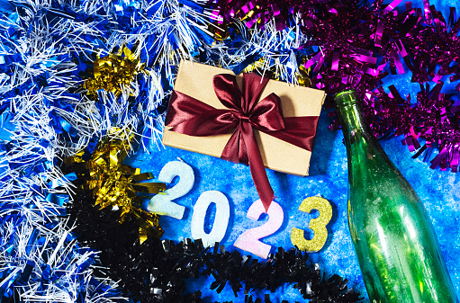 happy new year 2023 background new year holidays card with bright lights, tinsel and bottle and gift box on blue wood table