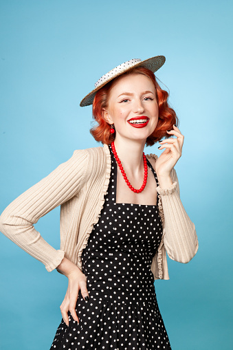 Studio portrait of a cheerful red-haired white woman dressed in retro style in a polka-dot dress against a blue background