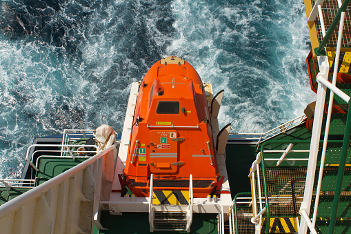 Top view on the orange colour life boat with reflective tape stickers secured with davits and safety hooks on the merchant cargo container vessel sailing through Adriatic Sea during sunny day.