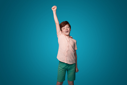 Smiling little boy raising hand up enjoying the win isolated on blue color wall background. Excited cheerful. Blue background. Happy family.