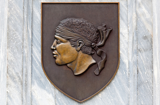 Cast bronze plaque of a Moors head with a bandana mounted on a shield background. This is the heraldic symbol of the French island of Corsica in the Mediterranean Sea. It's origins are slightly gruesome. According to legend, it originates from the 13th century when a young Corsican woman named Diana was captured by Moorish slavers who planned to sell her to the slave market of Granada in Spain. Her fiancé Pablo managed to free her and a battle ensued between Corsicans and Moors, during which the Moorish leader Mansour Ben Ismaïl was beheaded. His severed head then became the symbol of Corsica in remembrance of the event. The motif can be found all over the island, on flags and on buildings.