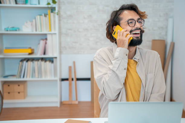 istock Young creative entrepreneur talking on the phone laughing smiling in his modern workspace at home. 1449796484
