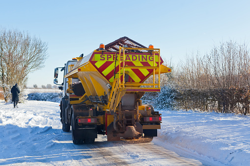 Treacherous snow and ice in sub-zero freezing conditions on rural UK countryside roads being gritted and salted by a public highways gritting vehicle to facilitate travel and help prevent accidents.