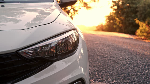 Close up view of front light of car headlights. unrecognizable white car is parked on side of road with setting sun in background. detail of exterior of modern car.
