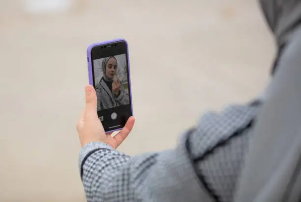 Photo of Young hijab woman doing makeup using phone screen.