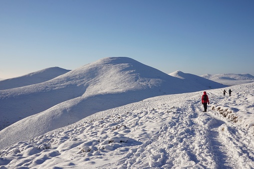 Pentland Hills, Scotland - 14 December 2022: Looking from a snowy path on Carnethy Hill in the Pentlands towards  more snow covered hills.  Three hillwalkers are wlaking on the snow covered trail.