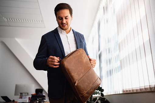 Young businessman at his workplace opening a leather backpack.