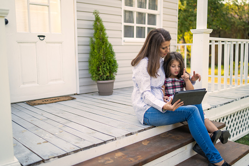 A little girl is sitting next to her mother on the front porch steps as they both look together at a digital tablet sharing family time
