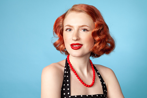 Studio portrait of a cheerful red-haired white woman dressed in retro style in a polka-dot dress against a blue background
