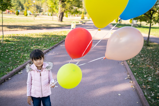 A small Asian girl wearing a winter coat is holding a bunch of five colorful balloons, standing on a footpath in a public park on a bright sunny day