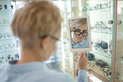 A woman, trying on new glasses and holding a mirror, in a bright modern opticians shop.