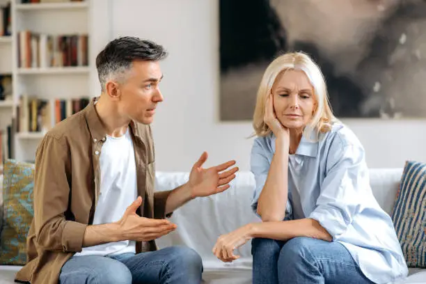 Emotional dialogue of a married couple in the living room on the couch. Outraged worried husband emotionally expresses his opinion to his wife, upset wife resting her head on her hand looking down