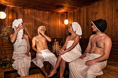 Four person with bath besoms sitting resting on bench in spa complex. Russian bathhouse. Two couple relaxing and sweating in wooden sauna, hot steam. Wellness, self care, healthy concept. Copy space