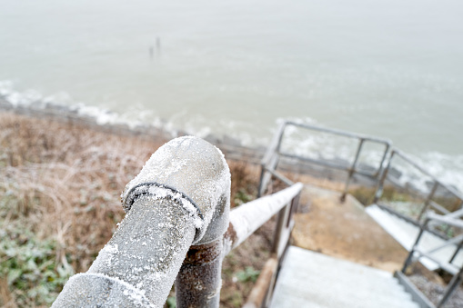 Shallow focus of a heavily frosted metal handrail located on a cliff-side staircase leading down to the beach. Treacherous in these icing conditions due to slipping.
