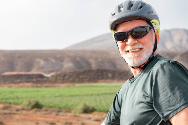 homem idoso ciclista feliz com capacete de proteção andando de bicicleta no campo em um dia ensolarado. bonito homem barbudo desfrutando de estilo de vida saudável e atividade esportiva - senior adult human face male action - fotografias e filmes do acervo