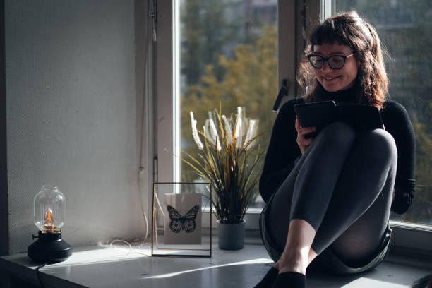 istock Young woman sitting on a window sill reading 1449844547